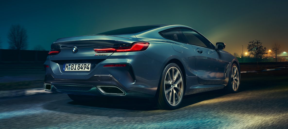 BMW M850i xDrive Coupé in Barcelona Blau metallic, Heckansicht, fahrend bei Nacht.